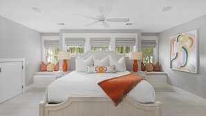 A bedroom with a large bed, two bedside tables, a ceiling fan, and a window with a view of trees. - Dual Primary Suites & Luxe Whimsical Interiors (Bluffton)
