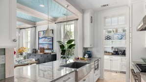 A modern kitchen with white cabinetry, a large island with a sink, and a view of the outdoors through large windows. - Dual Primary Suites & Luxe Whimsical Interiors (Bluffton)