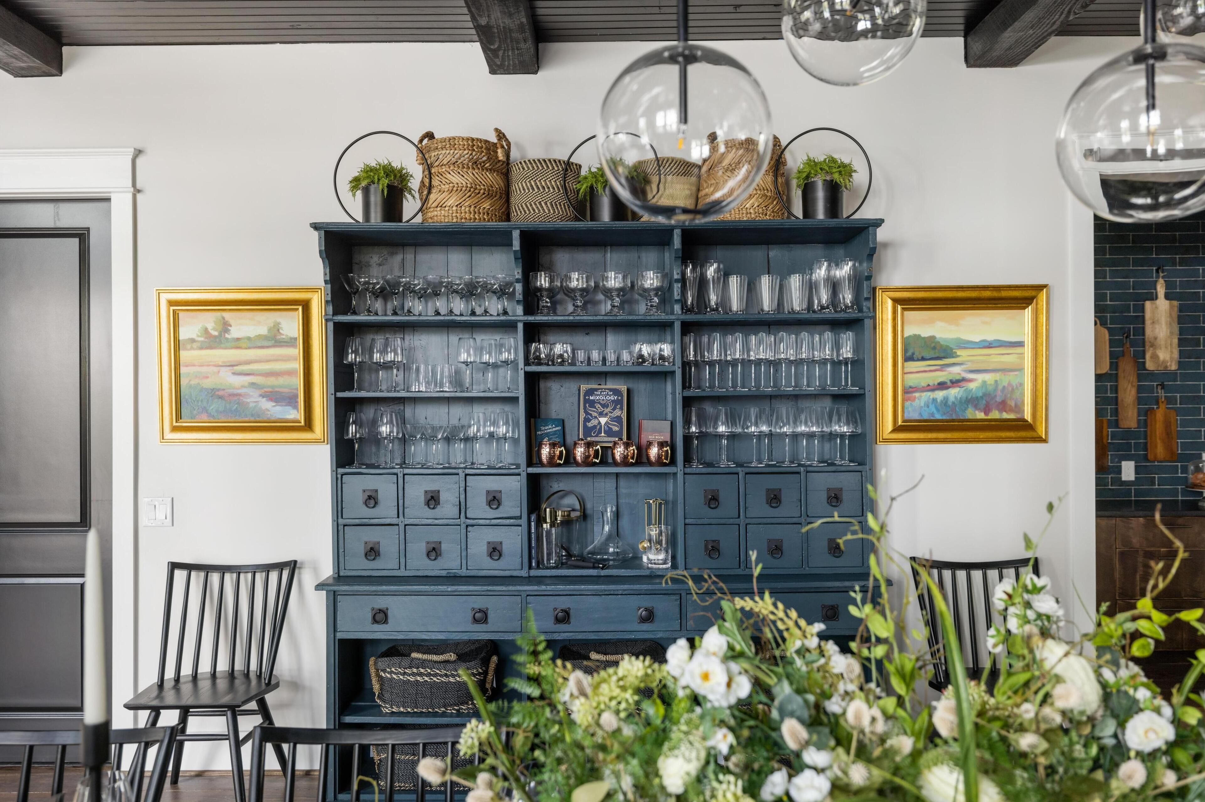 Een blauwe opslagunit met planken vol glaswerk en decoratieve items, omringd door stoelen en lijstwerken.