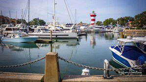 Un puerto deportivo con varias embarcaciones amarradas, un faro al fondo y una barrera de cadena en primer plano.