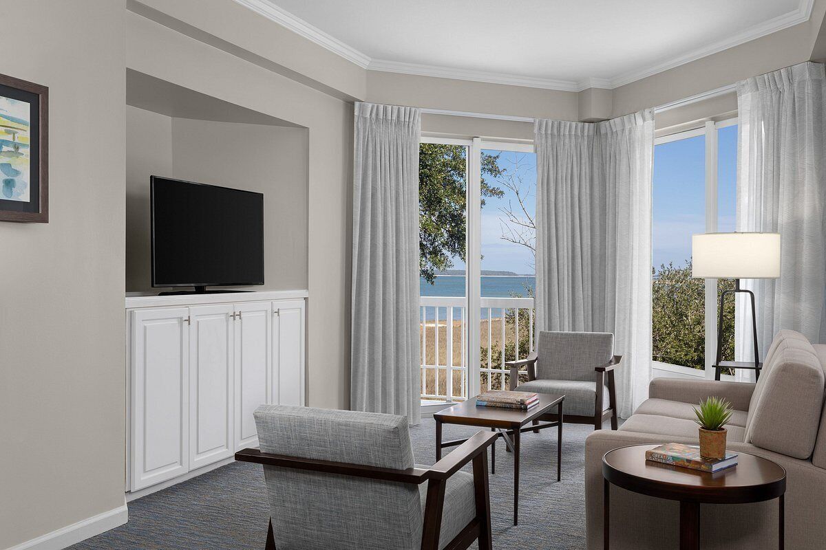 A hotel room with a flat-screen TV, a sofa, a chair, a small table, and a view of the outdoors.