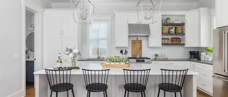 A modern kitchen with white cabinets, a central island with bar stools, a stainless steel refrigerator, and pendant lighting.