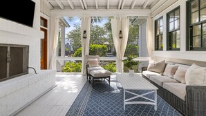 A covered porch with a white brick fireplace, a wicker sofa, and a dining table. - Luxe Retreat with Carriage House & Water Views (Bluffton)