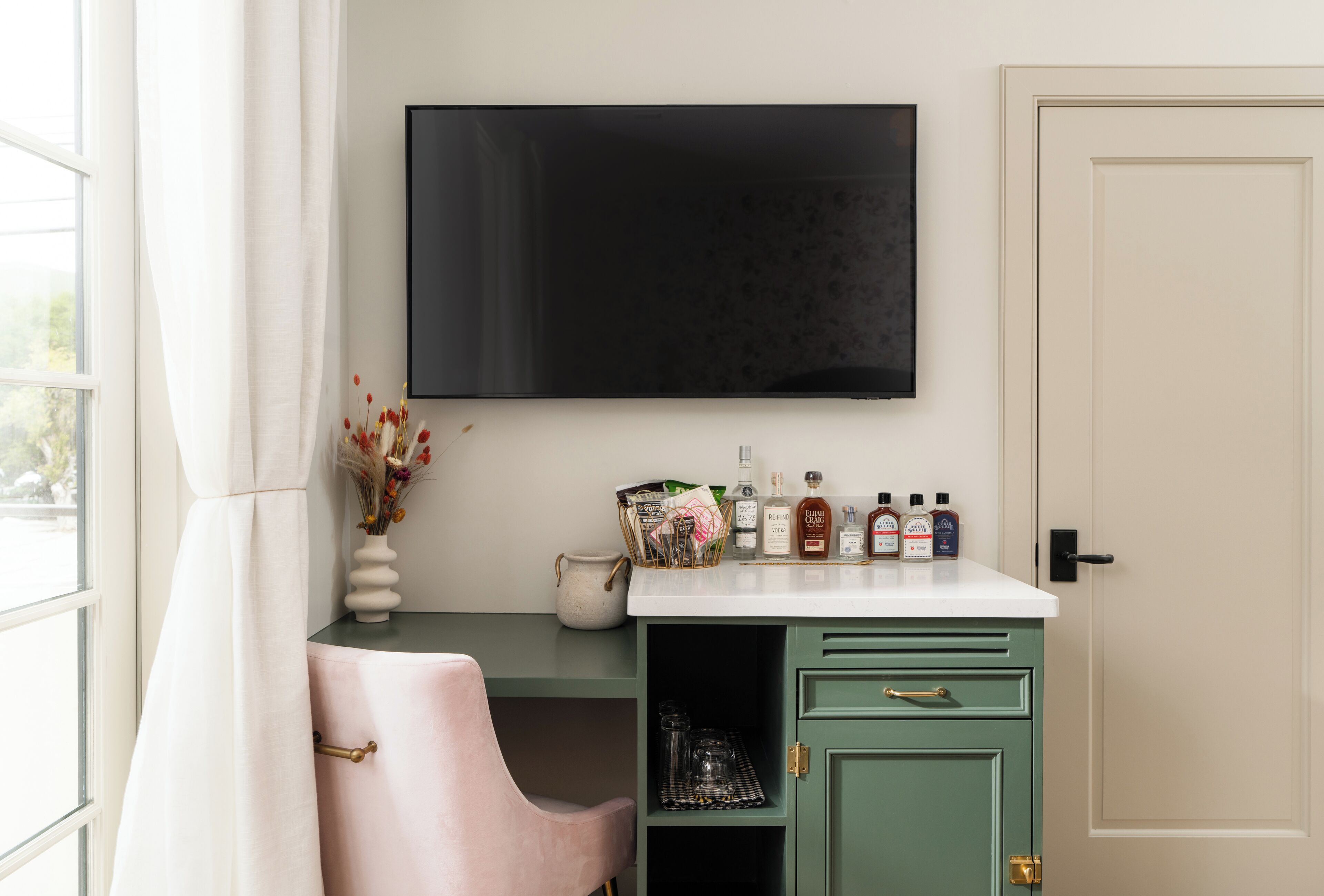 Una habitación con televisión montada en la pared, un escritorio con un florero de flores secas y una silla rosa.