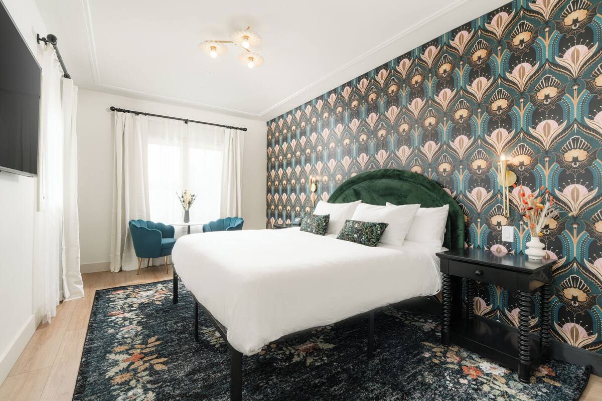 A bedroom with a large bed, a dark green headboard, a patterned wallpaper, a small side table, and a blue chair.