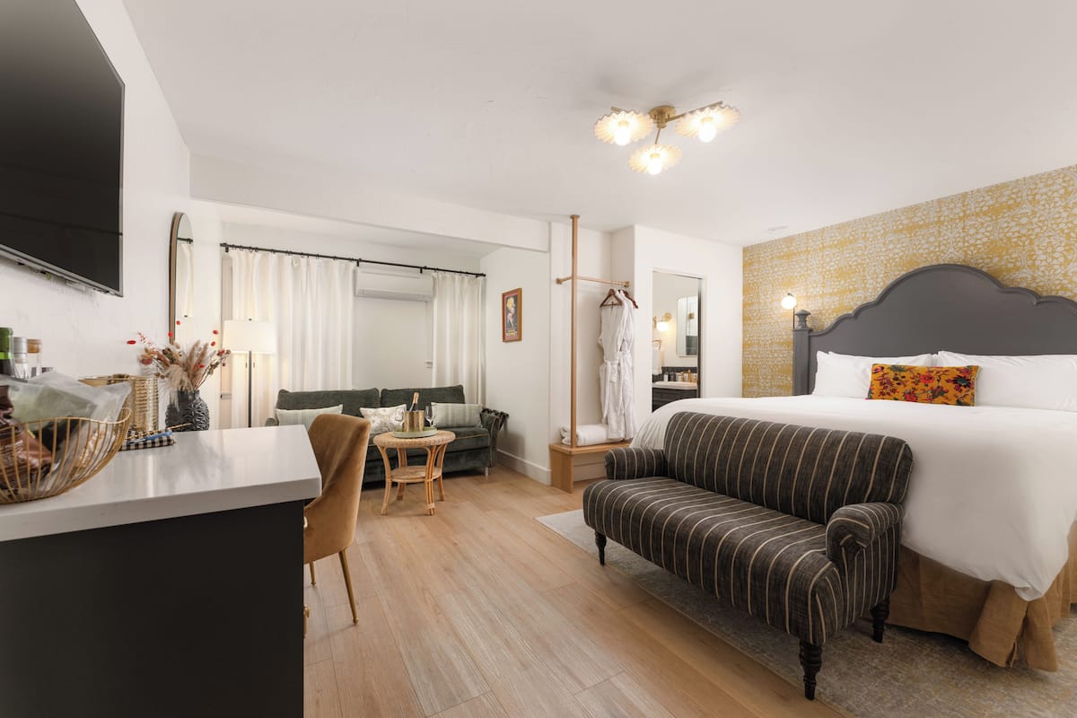 A hotel room with a bed, a sofa, a small table, a chair, a TV, and a mirror.
