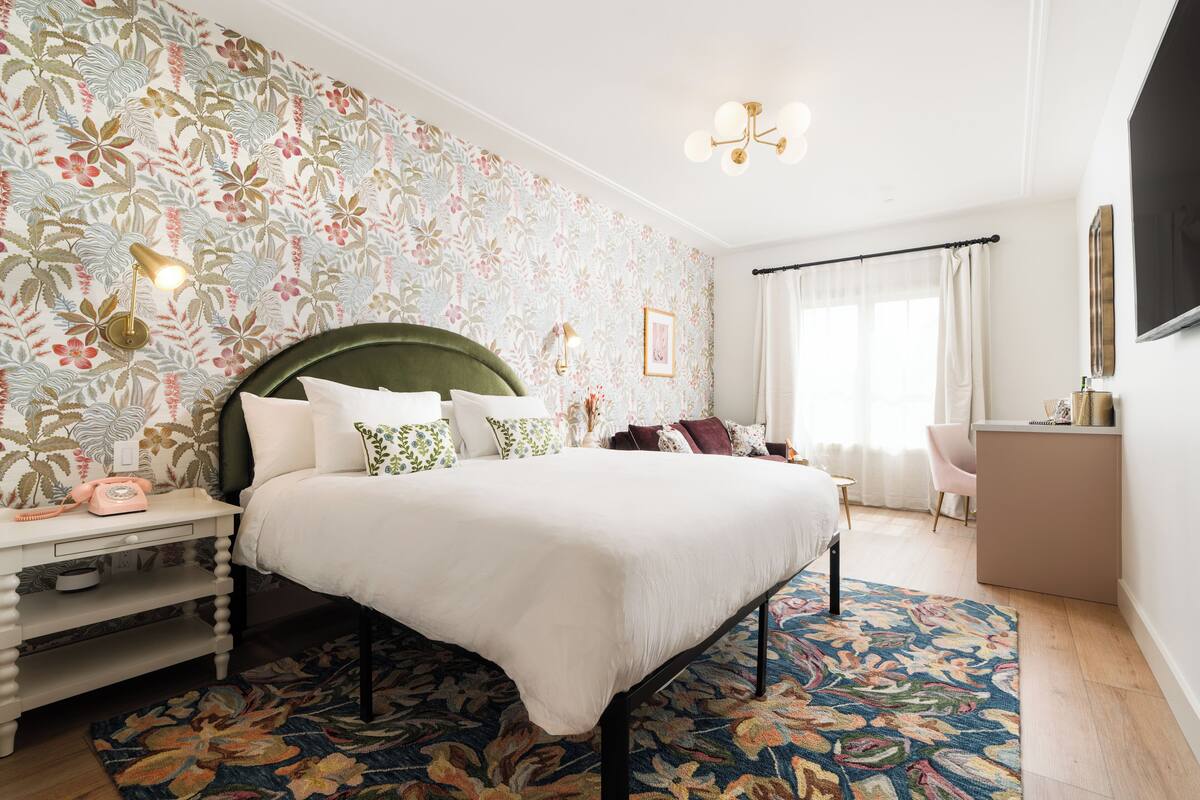 A bedroom with a floral wallpaper, a bed with white bedding, a nightstand with a telephone, and a desk with a chair.