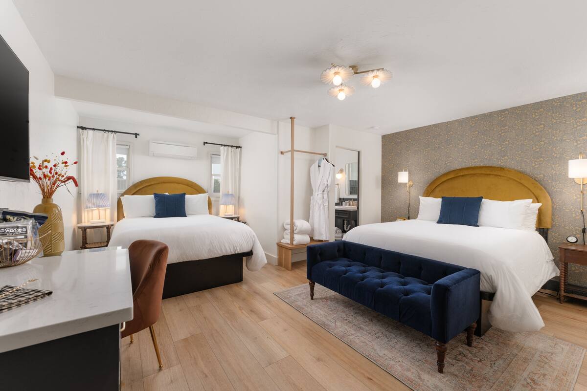 A hotel room with two beds, a desk, a chair, a bench, a TV, and a large mirror.