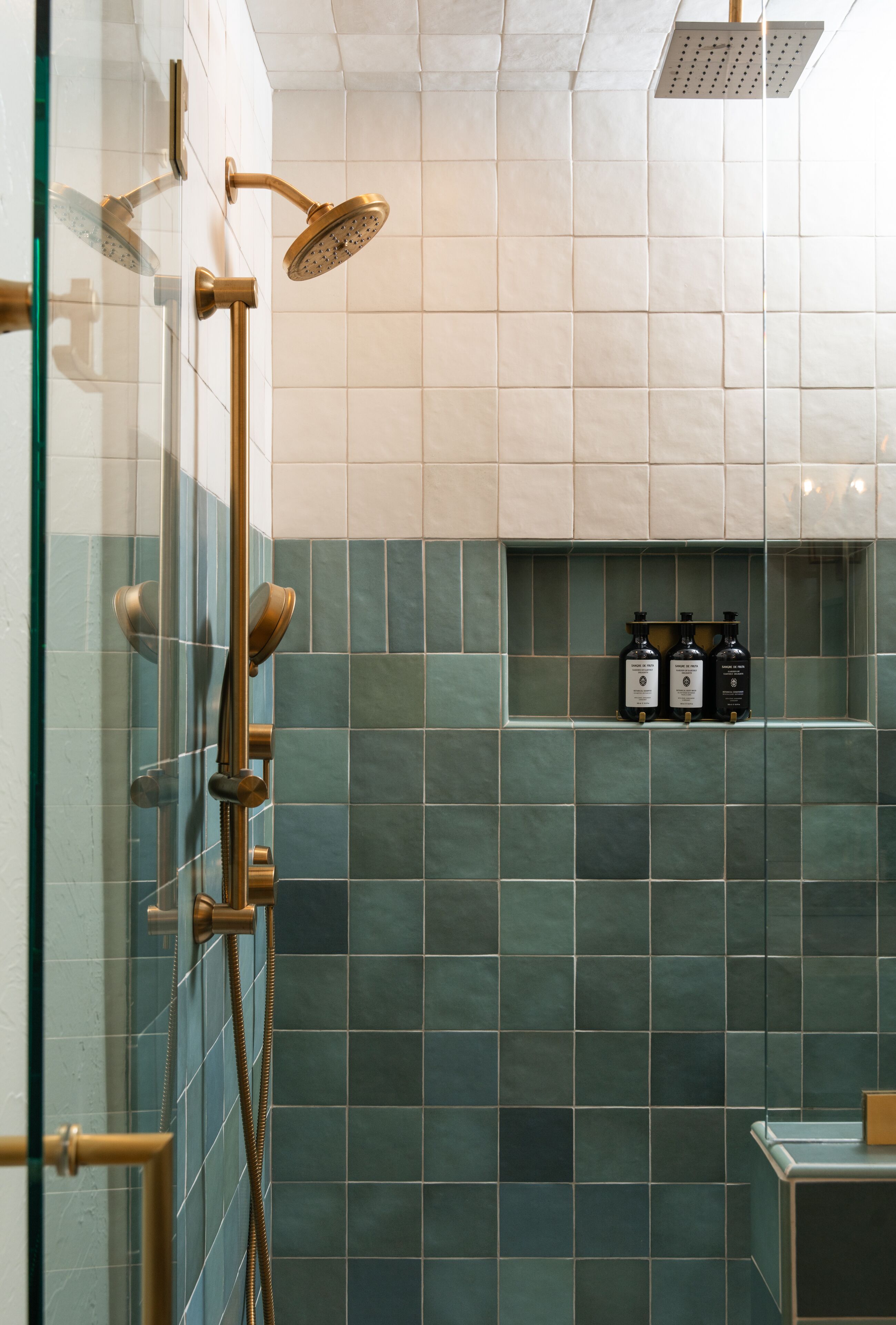 A modern bathroom with a glass shower enclosure, a handheld showerhead, and a wall-mounted faucet. There are three bottles of toiletries on a shelf above the tiled floor.