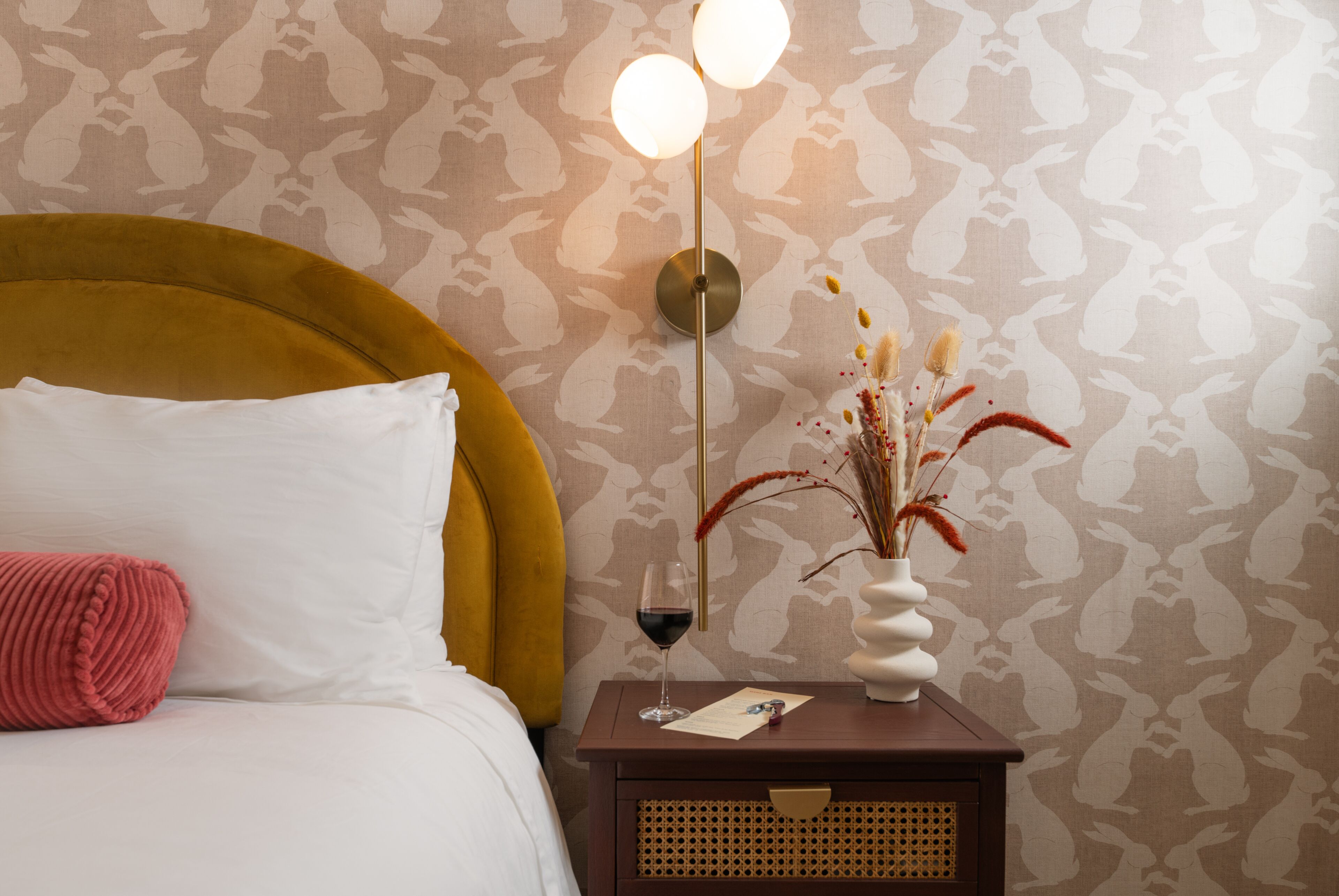 A neatly made bed with a white pillow and a red accent pillow, a bedside table with a vase of dried flowers and a glass of red wine, and a wall with a patterned wallpaper.