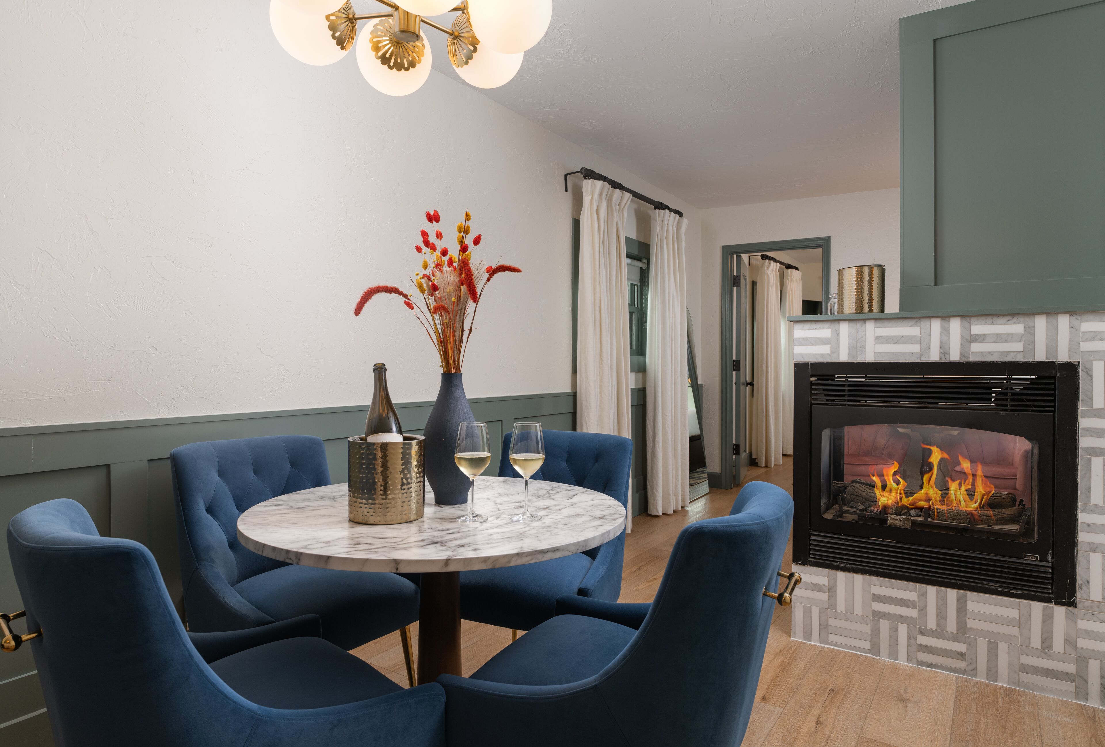 A dining area with a round table, blue upholstered chairs, a vase with flowers, and a fireplace with a visible fire.