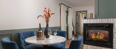 A dining area with a round table, blue upholstered chairs, a vase with flowers, and a fireplace with a visible fire.