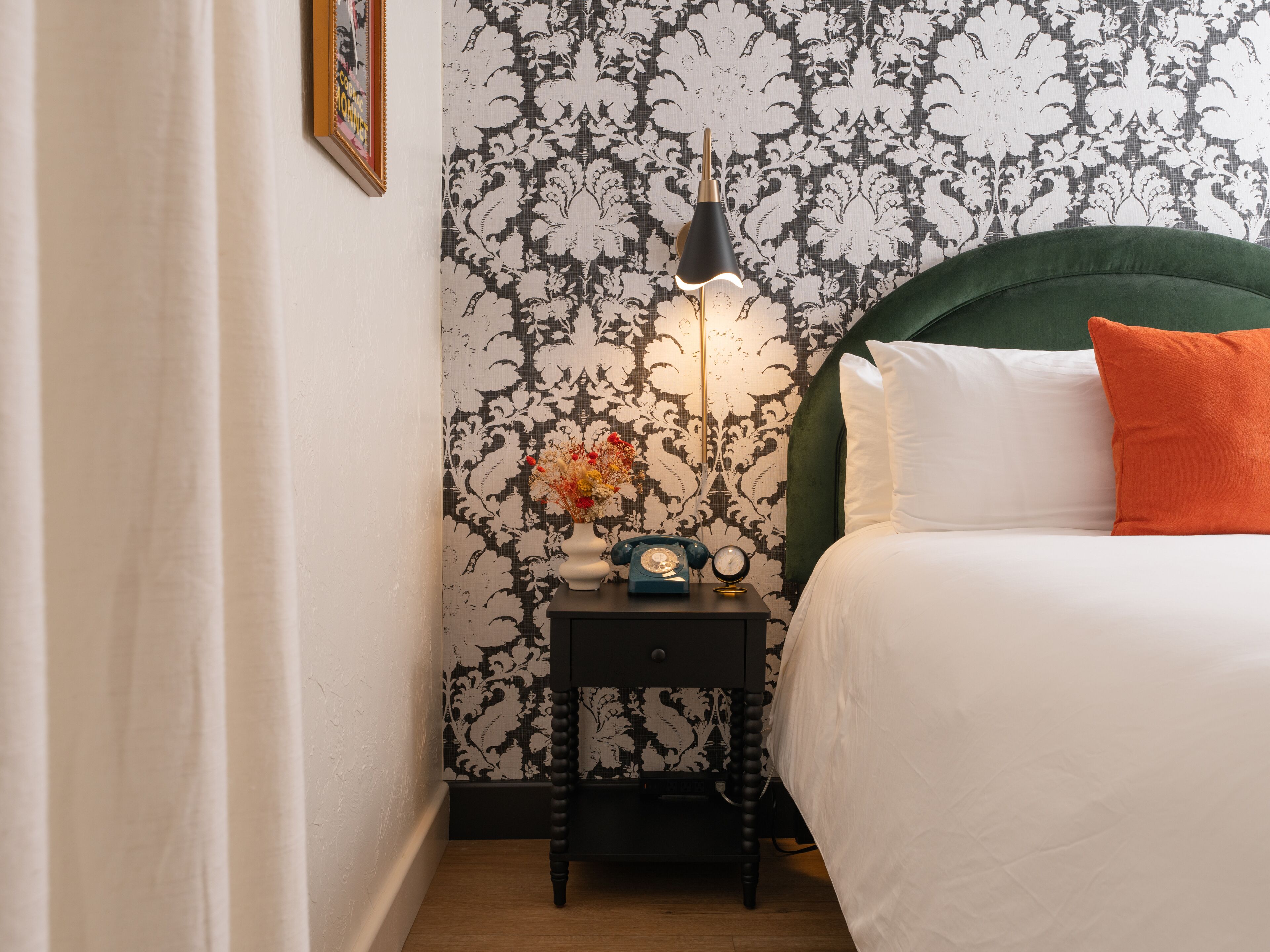 A neatly made bed with white and orange pillows, a green headboard, and a nightstand with a vintage telephone and a lamp.
