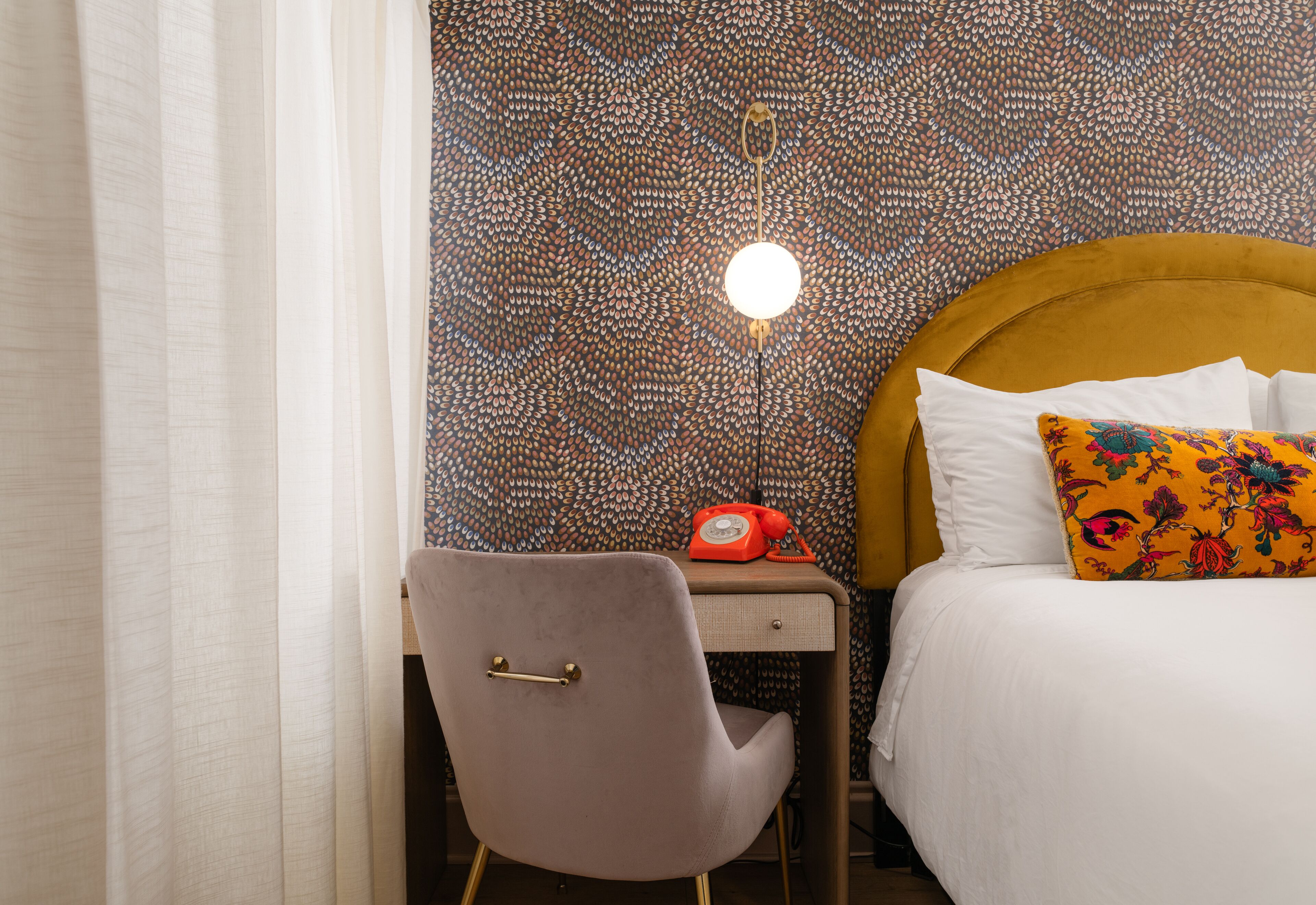 Una habitación de hotel con papel tapiz estampado, una cama con almohadas blancas y coloridas, un escritorio con teléfono y una silla.
