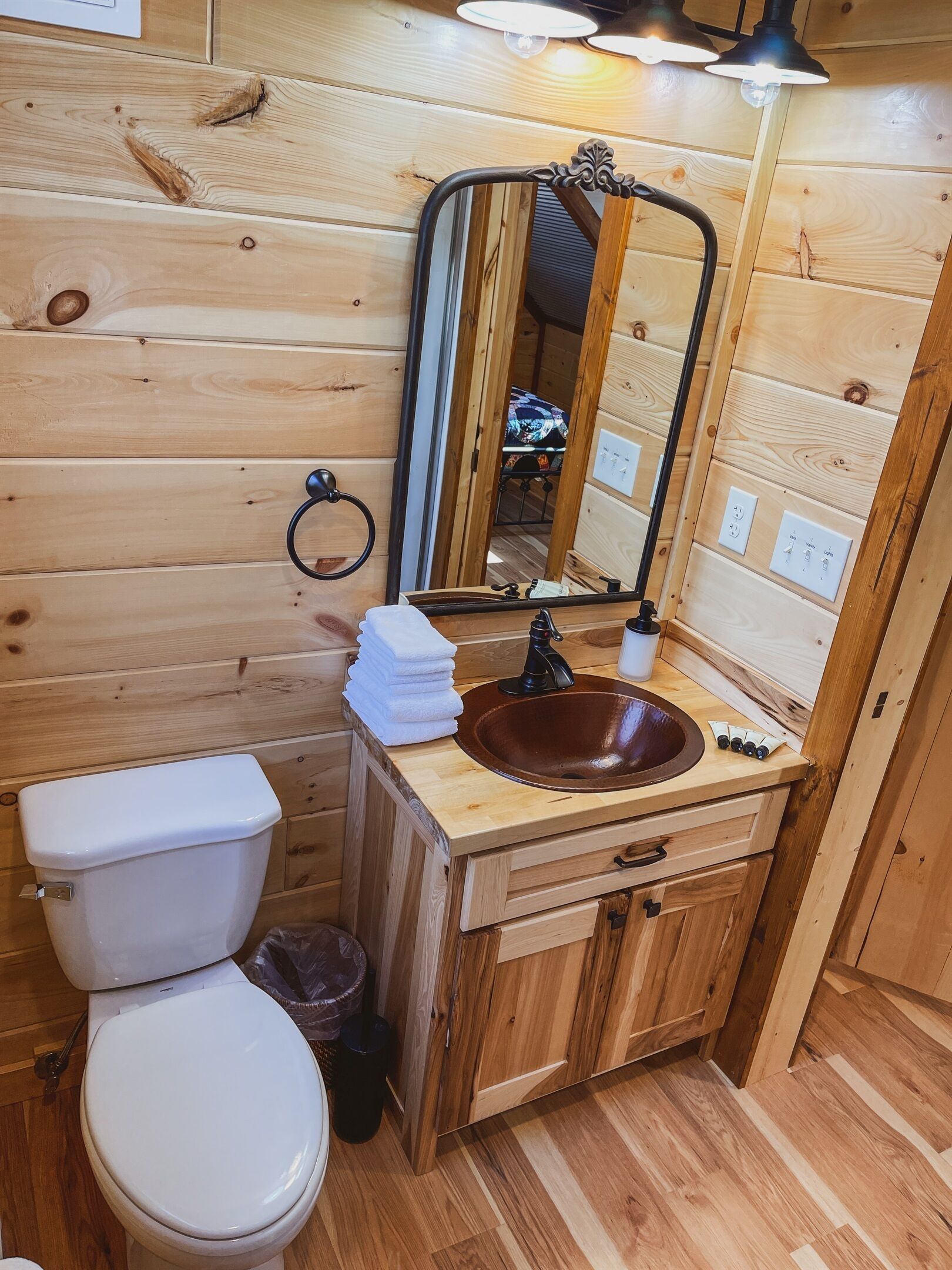 A wooden cabin bathroom with a white toilet, a wooden vanity with a brown sink, a mirror, and a small trash bin.