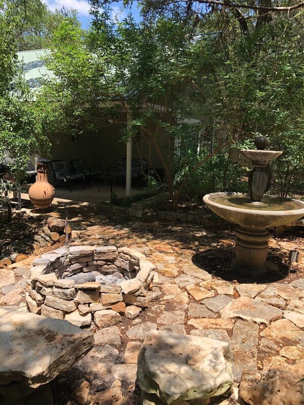 A stone fire pit surrounded by rocks, a large urn, and a birdbath in a garden area with trees and a covered seating area in the background. - Mystic Hill Suites (Wimberley)