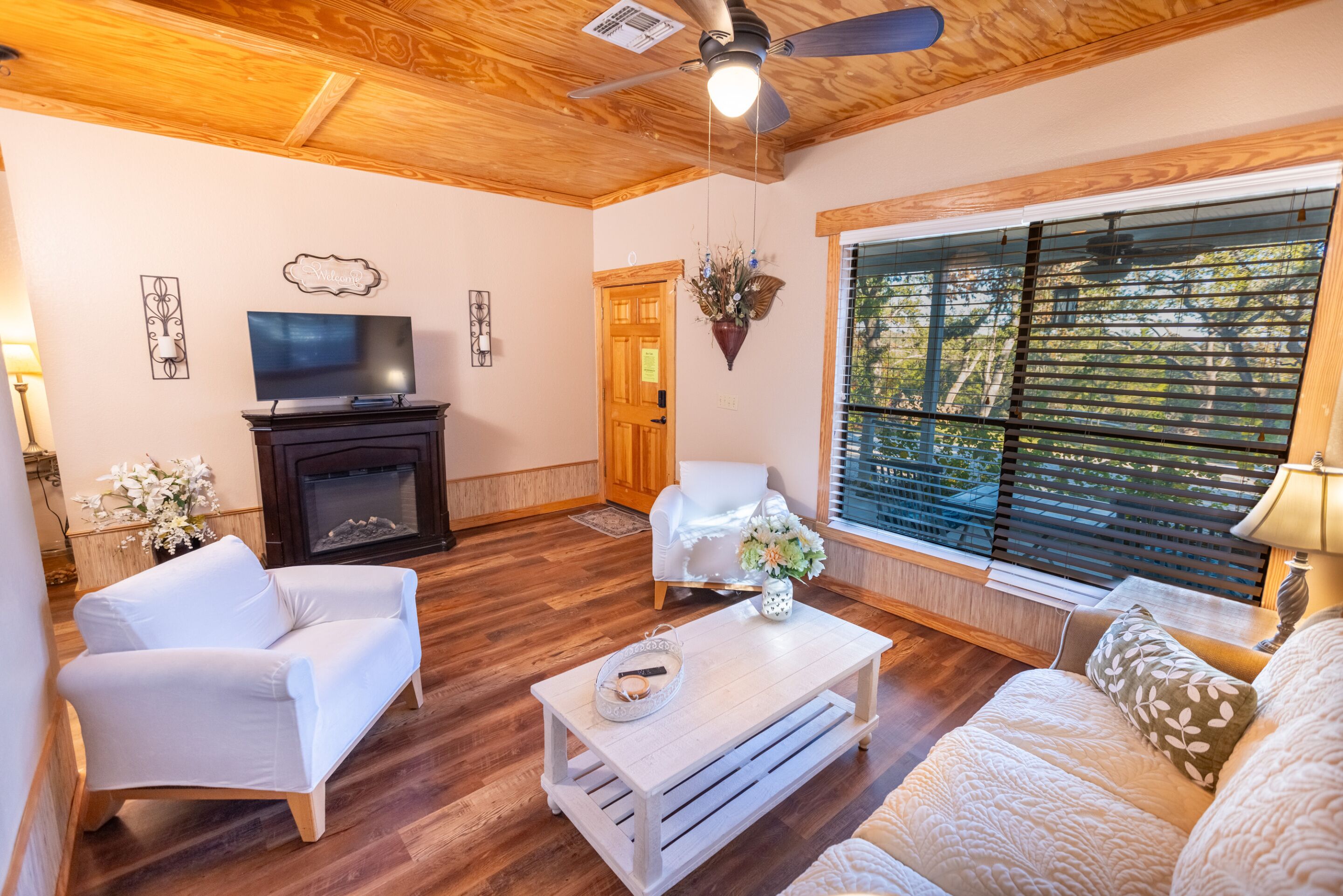 A living room with a fireplace, white sofas, a coffee table, and a large window with blinds.