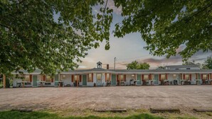 A motel with a central building and attached units, a gravel parking area, and trees framing the scene. - Sunset Acres Motel - Number 8 (Hudson)