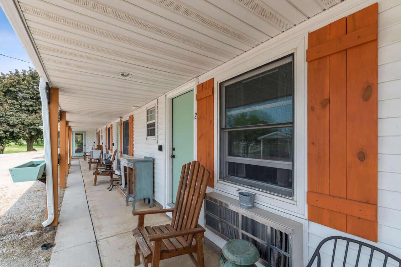 A row of motel rooms with wooden shutters, a covered porch, and a row of chairs.
