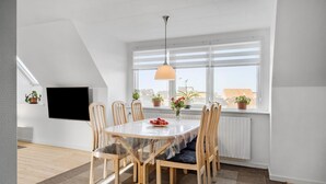 A dining area with a round table, six wooden chairs, a glass table cover, a hanging pendant light, and potted plants on the windowsill.