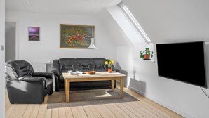 A modern living room with a black leather sofa, a wooden coffee table, a painting, and a large TV on the wall.