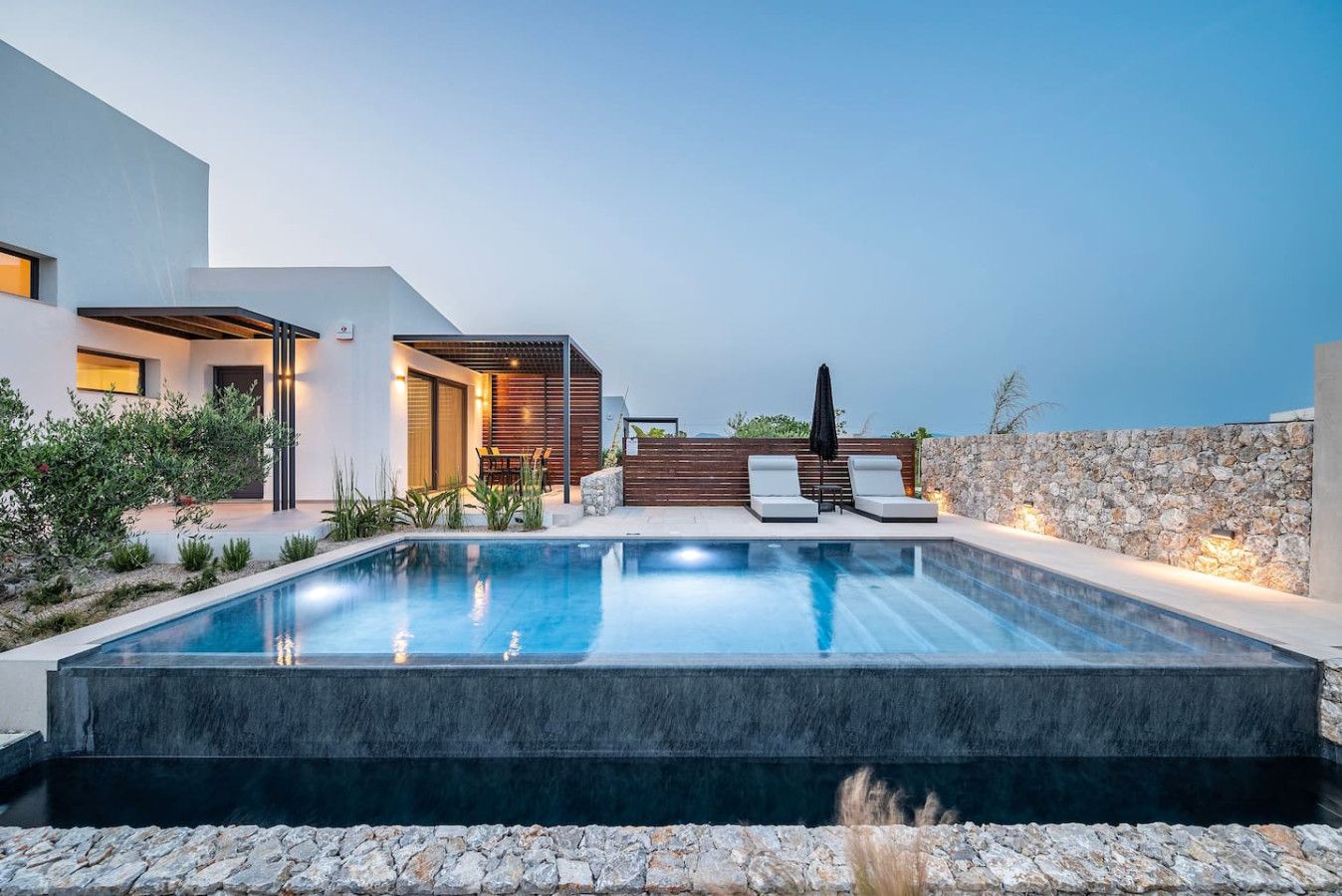 A modern outdoor pool area with a stone wall, lounge chairs, and a wooden deck.