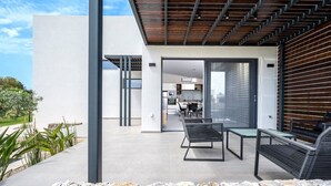 Une terrasse extérieure moderne, bordée de pierres, avec un espace repas comprenant une table et des chaises, et une cuisine visible à travers une porte ouverte.