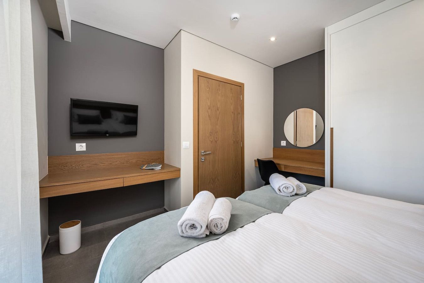 A modern hotel room with a bed, a TV, a wooden desk, and a mirror.