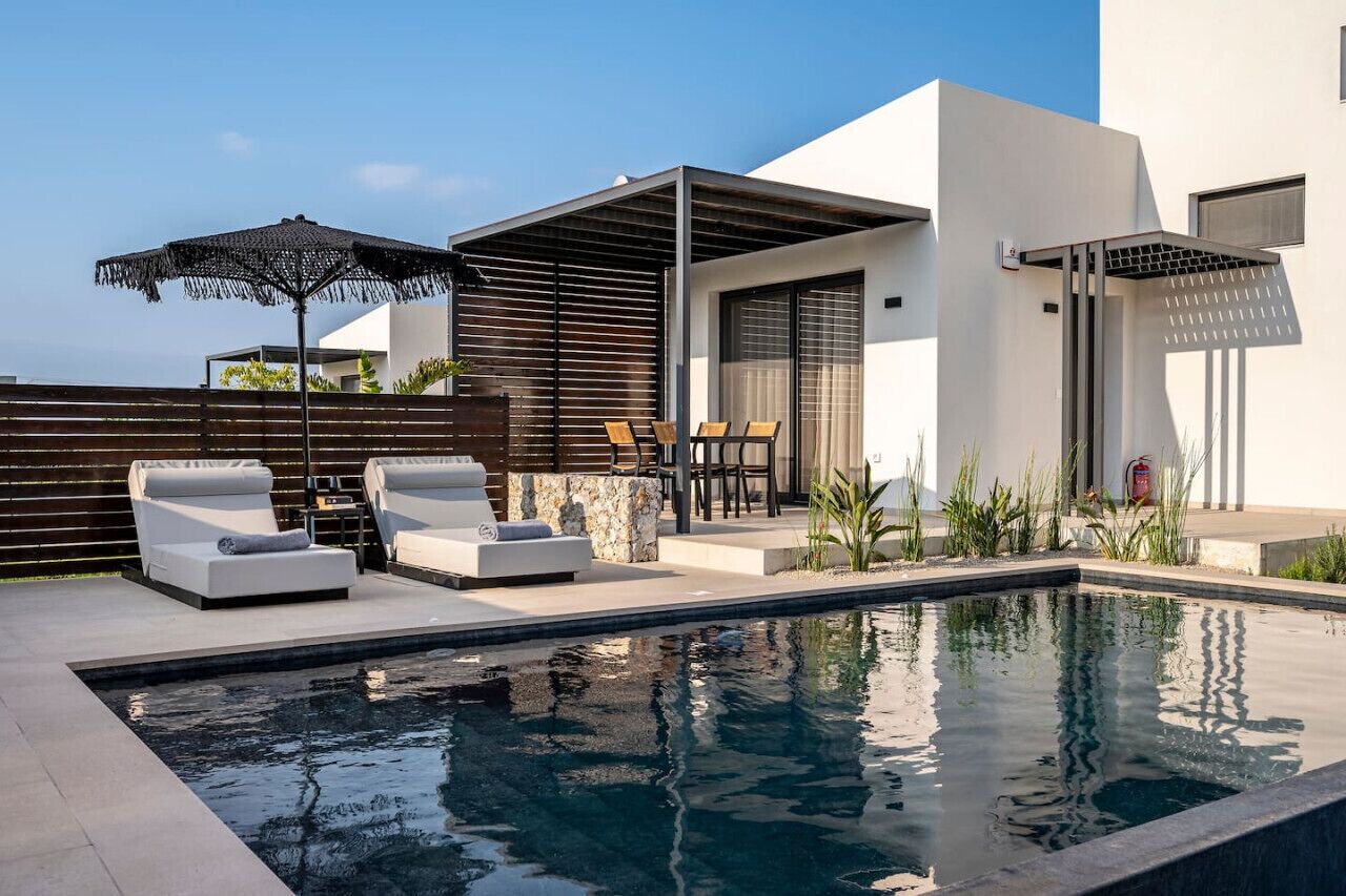 Et moderne poolområde udendørs med liggestole og parasoller, et overdækket opholdsområde samt et minimalistisk arkitektonisk design.