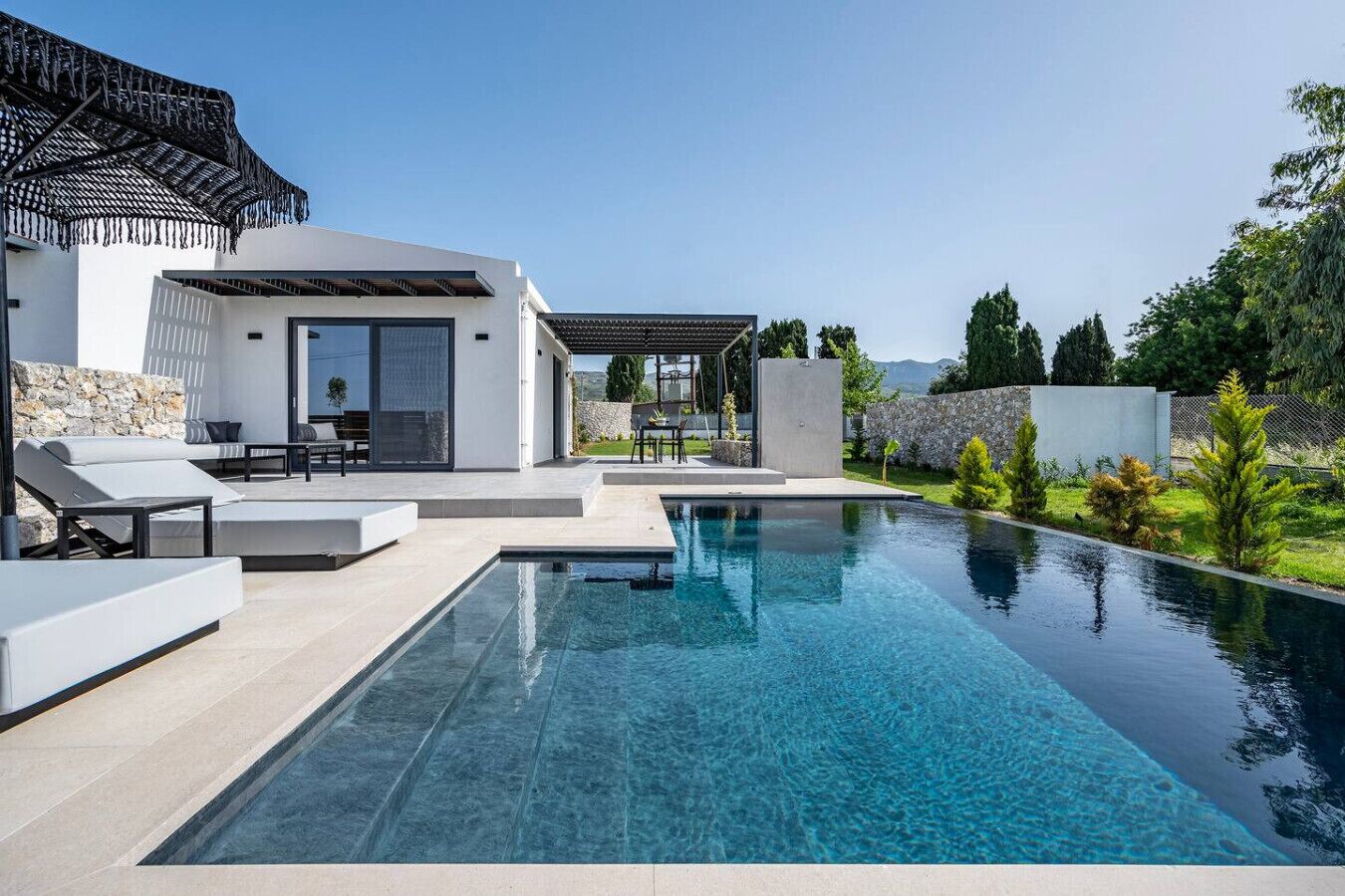 A modern outdoor pool area with a pergola, lounge chairs, and a stone wall.