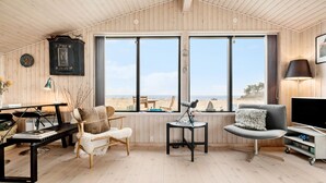 Een knus ingerichte kamer met houten wanden en vloer, grote ramen met uitzicht op een strand, een telescoop, een comfortabele bank en een televisie.