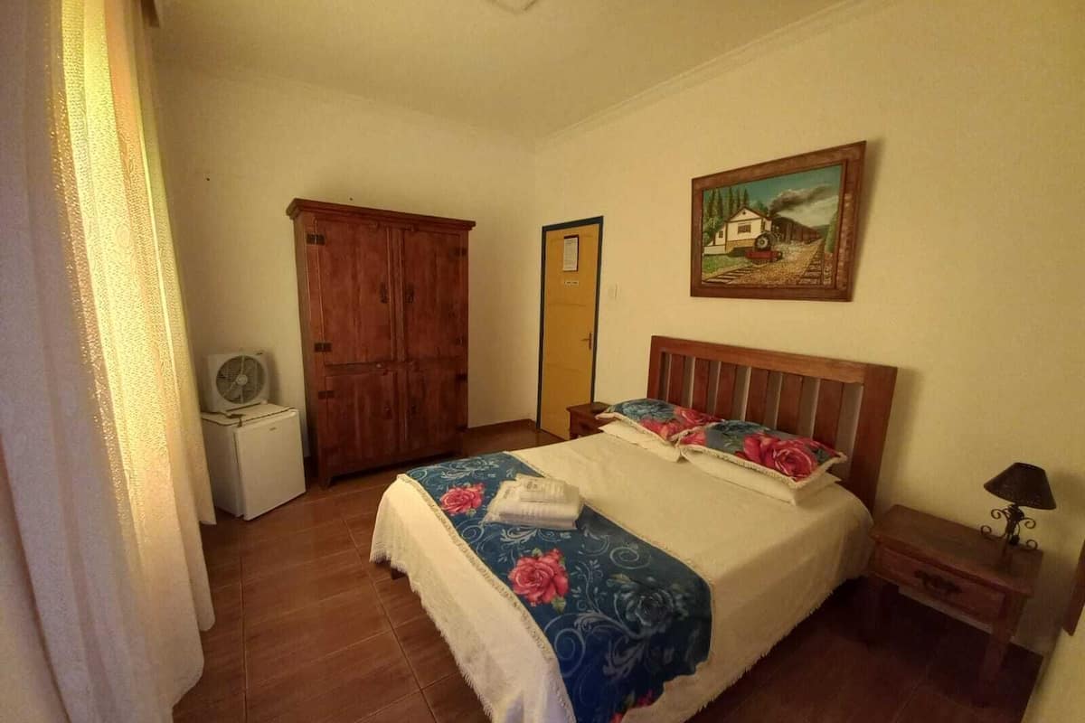 Quarto com cama de madeira, mesa de cabeceira, pequena mesa, guarda-roupa e um quadro na parede.
