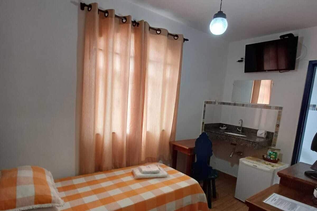 Quarto pequeno e arrumado, com cama, escrivaninha e cozinha compacta.