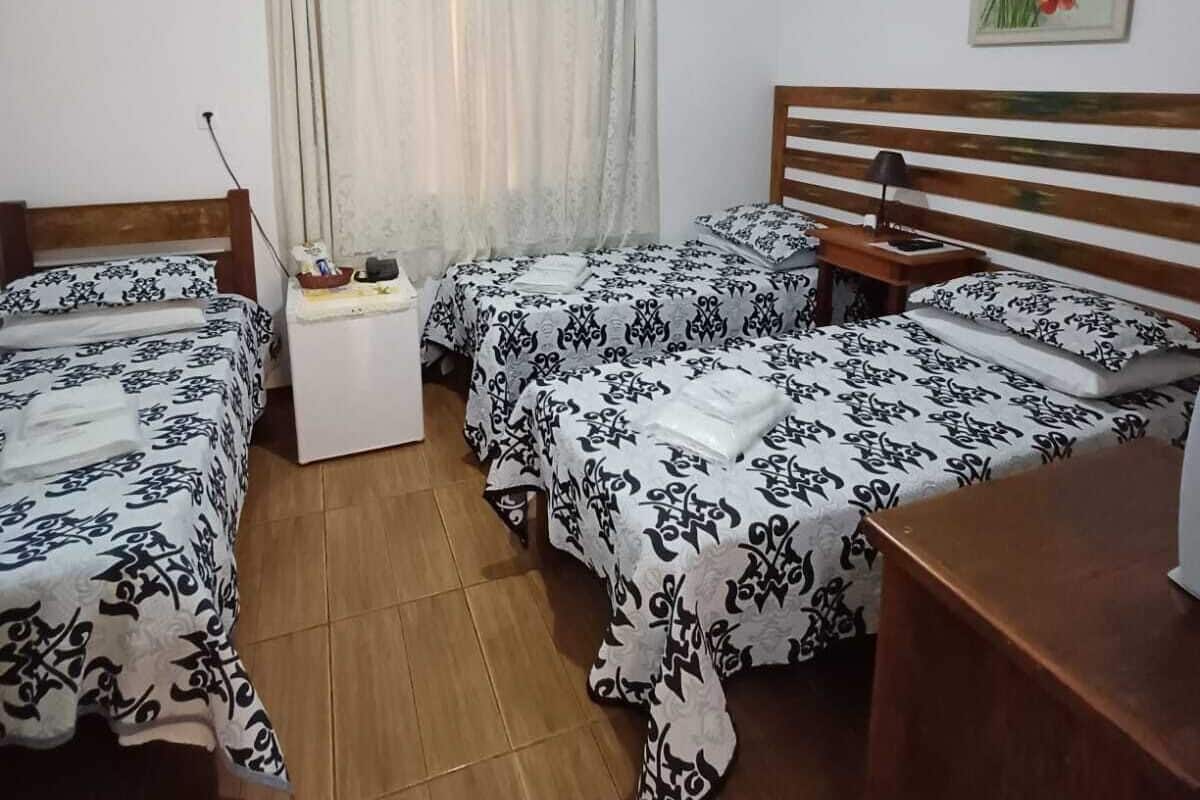 Quarto com duas camas, uma mesa pequena, mesa de cabeceira, luz no teto, janela com cortinas e um quadro na parede.