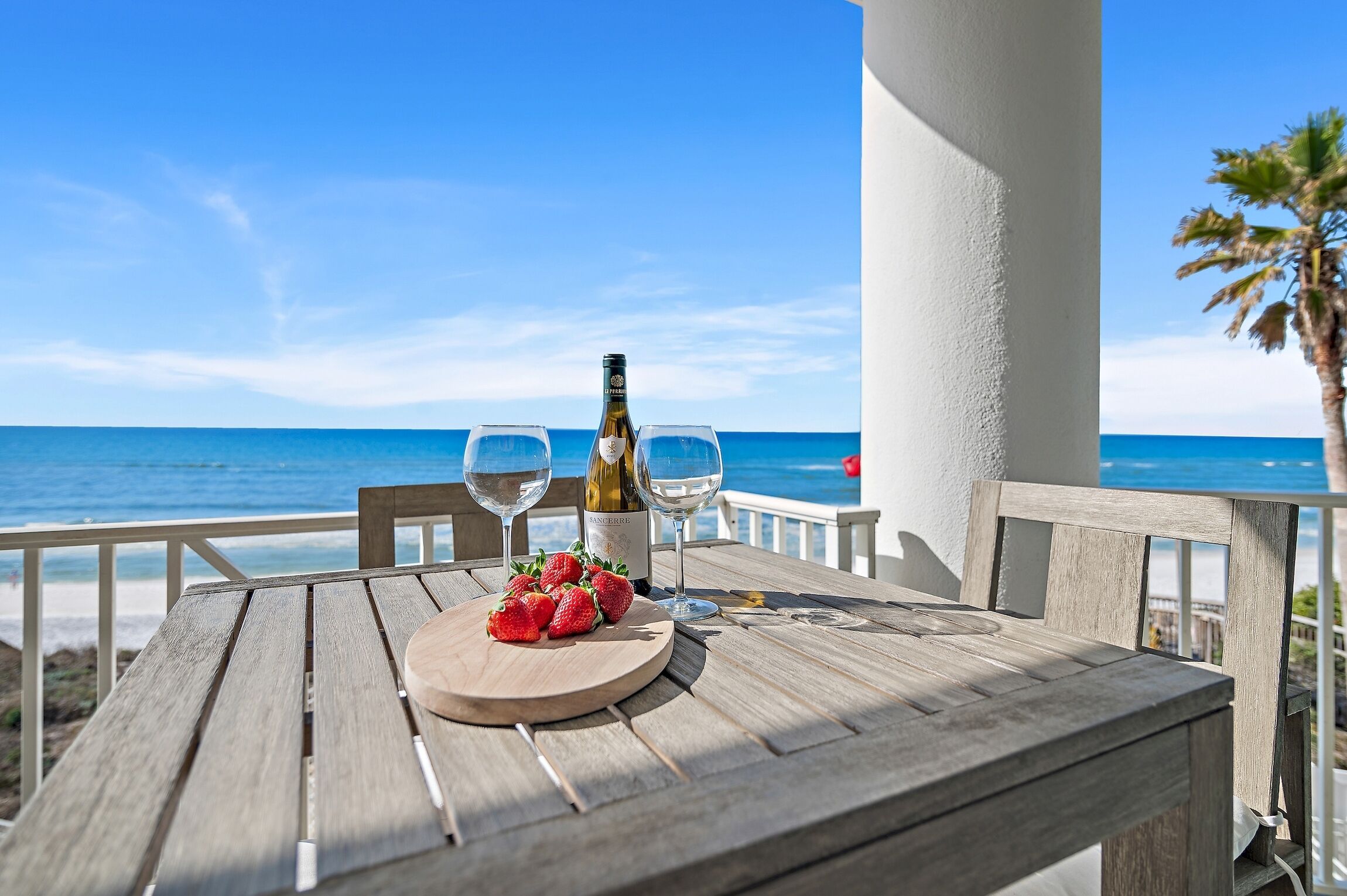 A wooden table with a bottle of wine, two glasses, and strawberries on a balcony overlooking the sea.