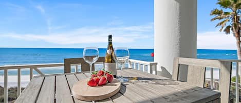A wooden table with a bottle of wine, two glasses, and strawberries on a balcony overlooking the sea.