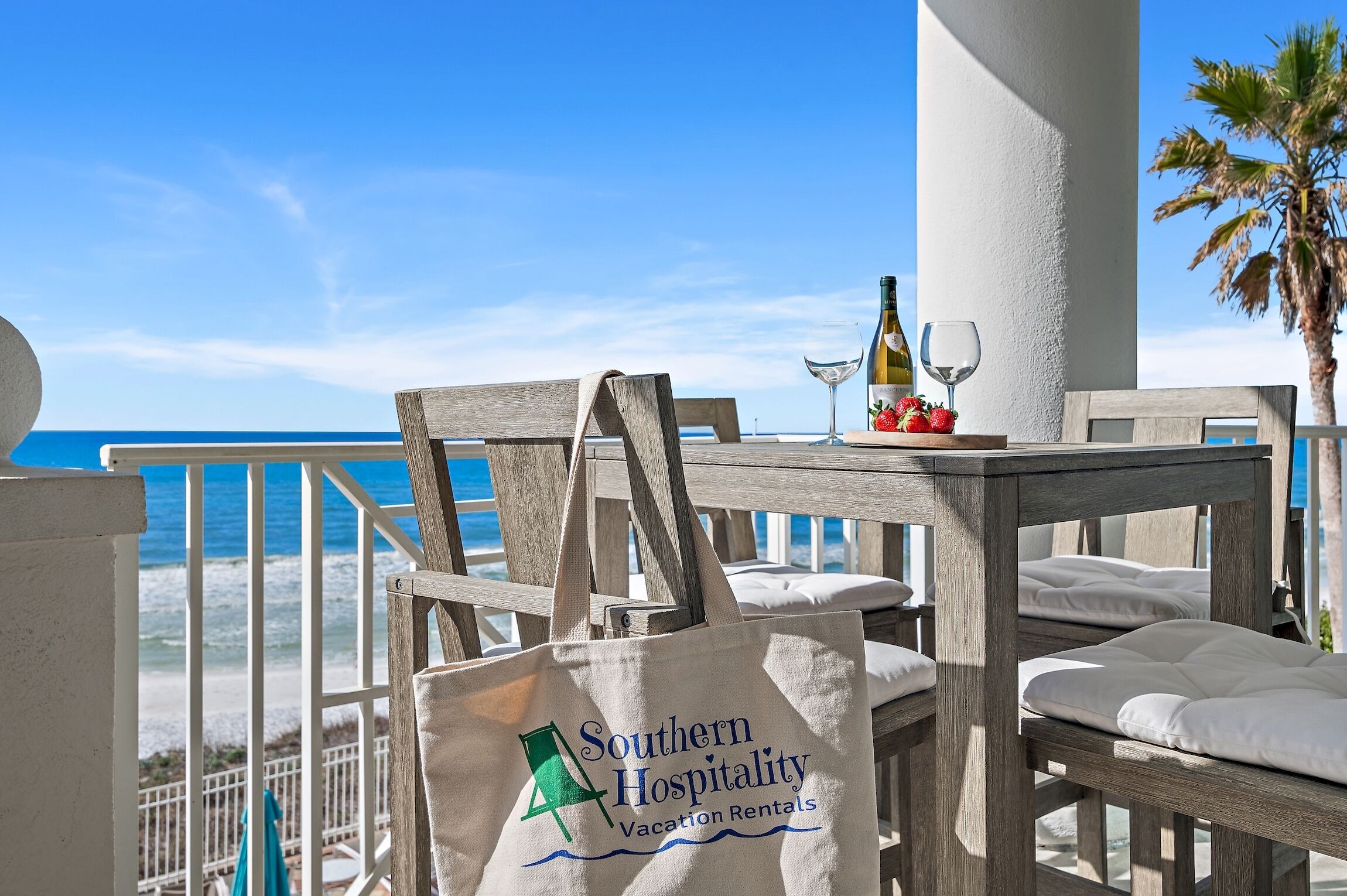 Une table en bois avec une bouteille de vin et un verre, un sac fourre-tout orné d’un logo, et une vue sur l’océan.