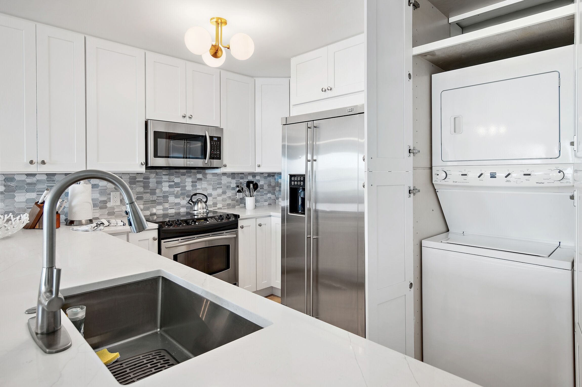 Una cocina moderna con armarios blancos, electrodomésticos de acero inoxidable, microondas integrado, refrigerador de acero inoxidable y un lavamanos con grifo.