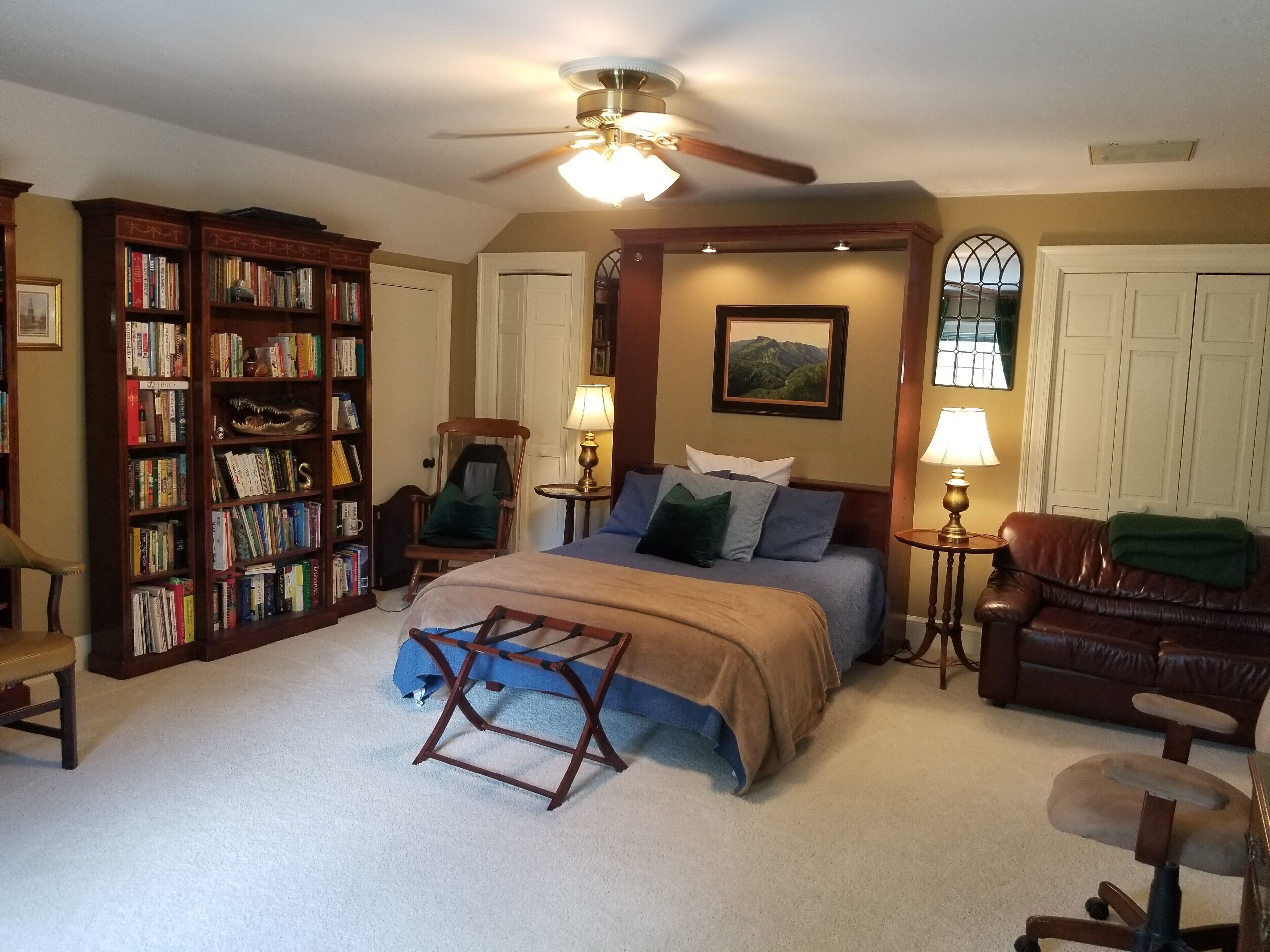 A bedroom with a large bed, a bookshelf, a desk, a chair, and a ceiling fan.