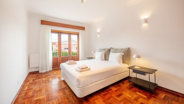 Un dormitorio con piso de madera, una cama con ropa de cama blanca, una mesita de noche con una lámpara y una ventana con cortinas.