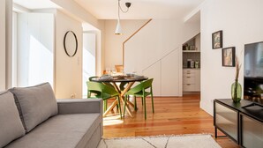 A modern living room with a grey sofa, a dining area with a round table and green chairs, a kitchen with white cabinets, and a TV on a black stand. - JOIVY Splendid downtown penthouse (Lisbon)