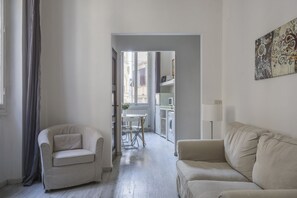 Un salon comprenant un canapé, un fauteuil, une table à manger avec des chaises, et un coin cuisine visible à travers une porte ouverte.