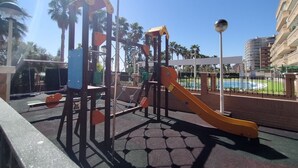 A playground with a slide and climbing structures, surrounded by palm trees and a pool. - ACV - Vistamar Iii-1ª Linea Planta 2 Norte 2 (OROPESA)