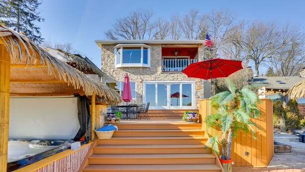 A wooden deck leading to a stone house with a balcony, a hot tub, and outdoor furniture. - Greenwood Lake Cottage w/ Shared Dock & Hot Tub! (Hewitt)