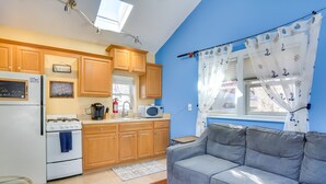 A compact kitchen with wooden cabinets, a white stove, and a microwave. There's a gray sofa and a window with curtains. - Greenwood Lake Cottage w/ Shared Dock & Hot Tub! (Hewitt)