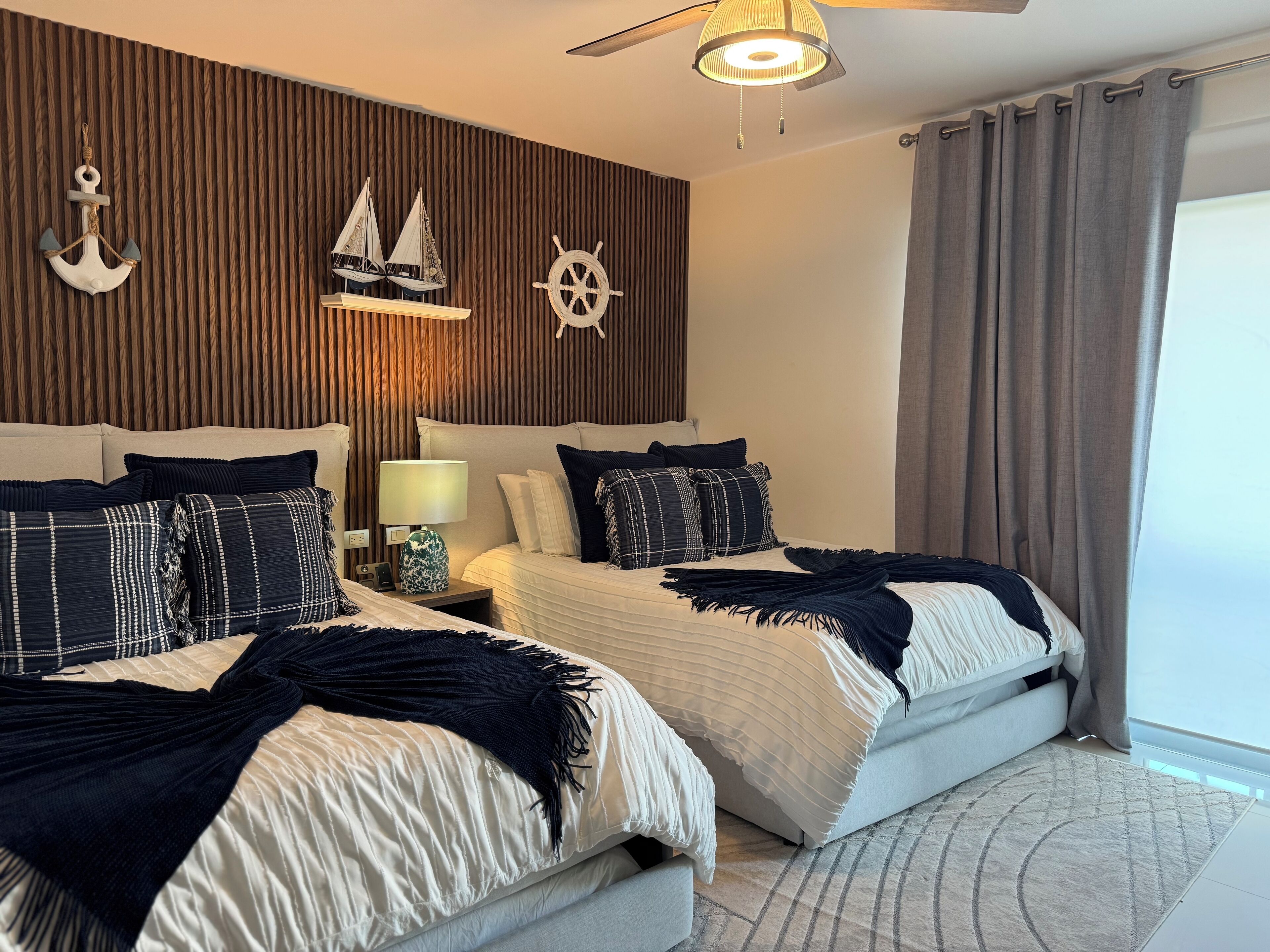 A modern bedroom with two beds, a wooden wall panel, a ship's wheel decoration, and a ceiling fan.