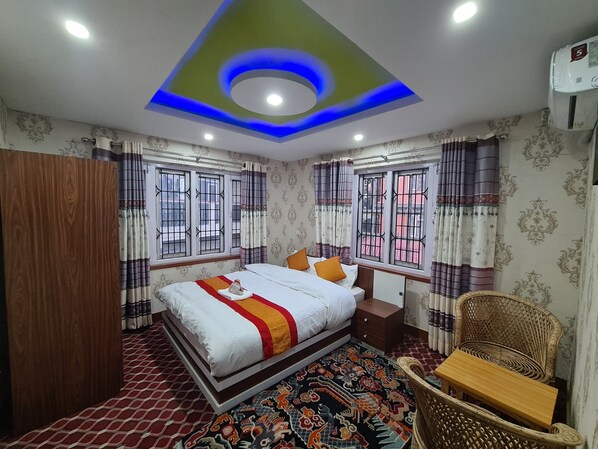 Habitación de hotel con cama, armario de madera, una silla de mimbre, una mesita de madera, cortinas estampadas y techo decorativo.