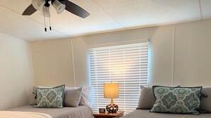 A bedroom with two beds, a ceiling fan, a bedside table with a lamp, and a window with blinds.