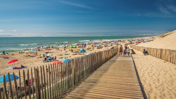 A beach with a wooden boardwalk leading to the shore, sandy beach, and umbrellas. - Proche Lac & Océan | Piscine | Camping 4* (Hourtin)