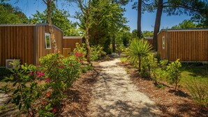 A pathway leading to wooden cabins surrounded by lush greenery and flowering plants. - Proche Lac & Océan | Piscine | Camping 4* (Hourtin)